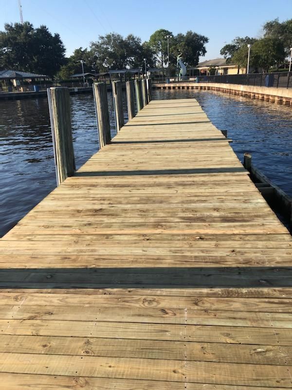 Wooden dock extends over water, with pilings on either side. Clear blue sky, trees in the background.