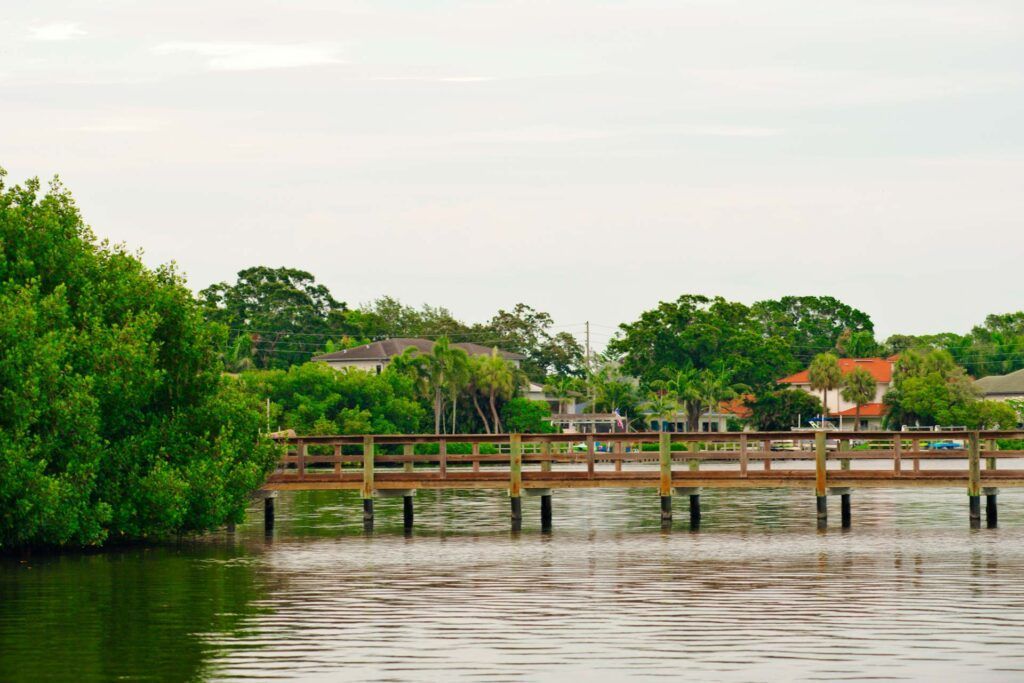 Wooden footbridge over calm water, surrounded by green foliage and houses under a cloudy sky.
