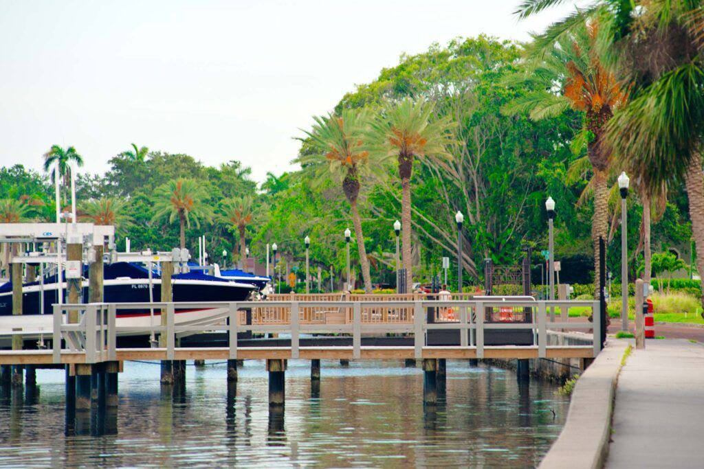 Wooden pier with boats, palm trees, and a walkway along water. Lush green trees in the background.