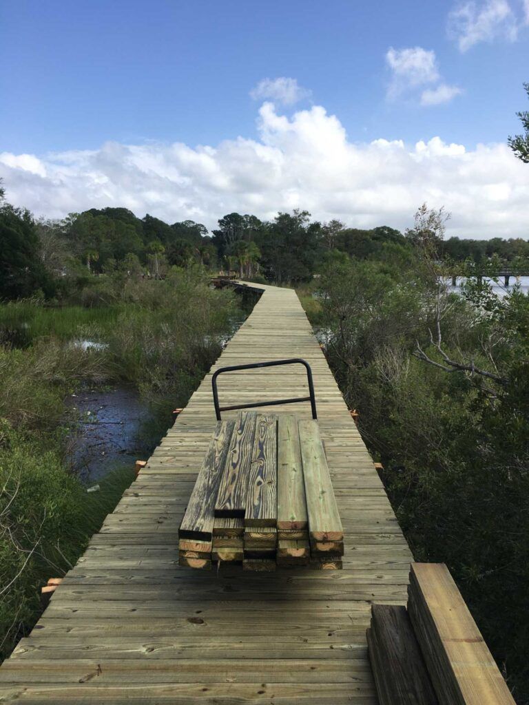 Wooden boardwalk in a marsh, with a hand truck loaded with lumber. Blue sky and foliage in the background.