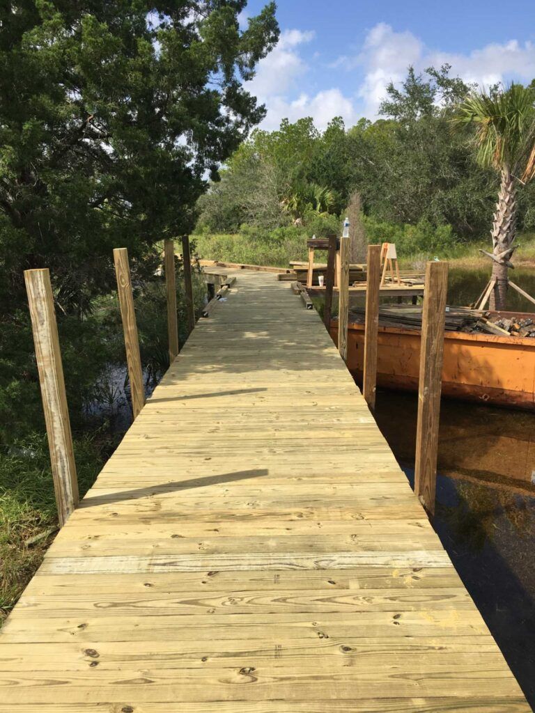 Wooden dock extending into water, with boats docked to the right, trees and blue sky background.