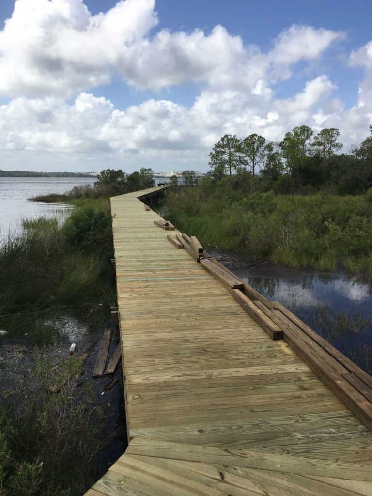 Wooden boardwalk curving along a marshy shoreline, under a partly cloudy sky.