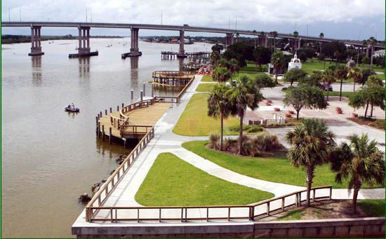 Waterfront park with bridge in background; wooden boardwalk, green grass, palm trees, and brown water.