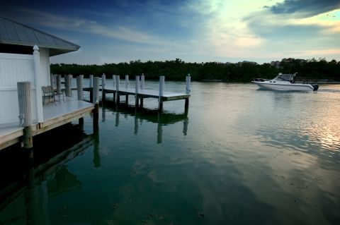 Dockside view with boat on calm water; white building, wooden dock, lush greenery, and cloudy sky.