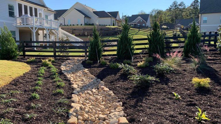 Landscape with a dry creek bed, trees, fence, and houses on a sunny day.