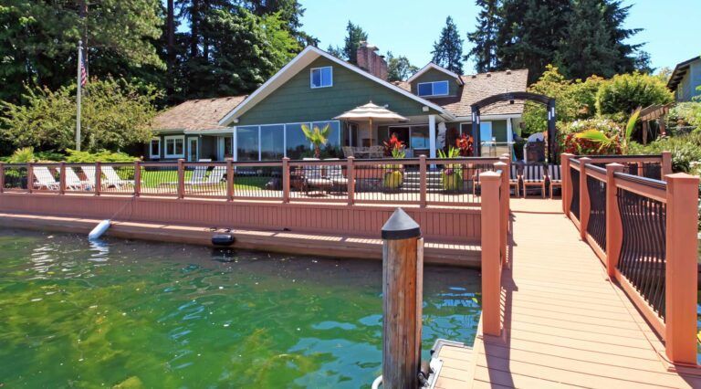 Lakeside green house with dock and patio. Sunny day, clear water, and trees in background.