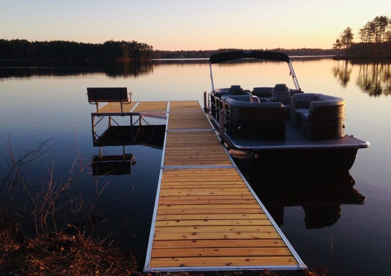 Pontoon boat docked at wooden pier on calm lake at sunset.