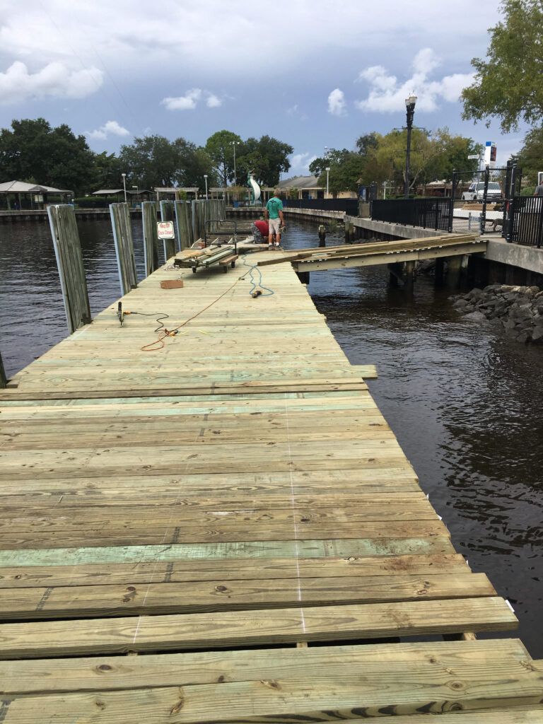 Wooden dock under repair with person, water, trees, and houses in the background.