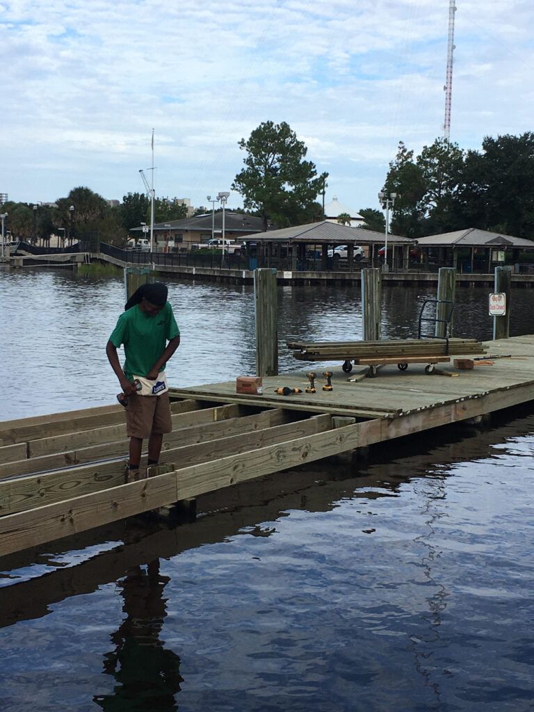 Person working on a wooden dock over water; green shirt, cloudy sky, buildings in the background.