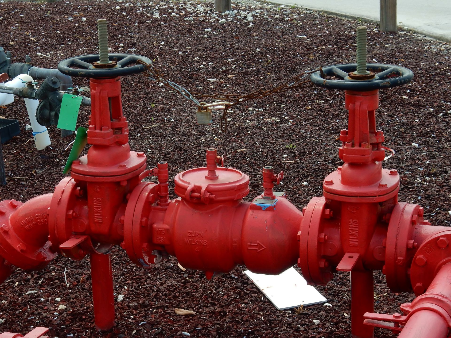 Red industrial pipeline valves and fittings on a gravel surface, with black handwheels and bright red paint.