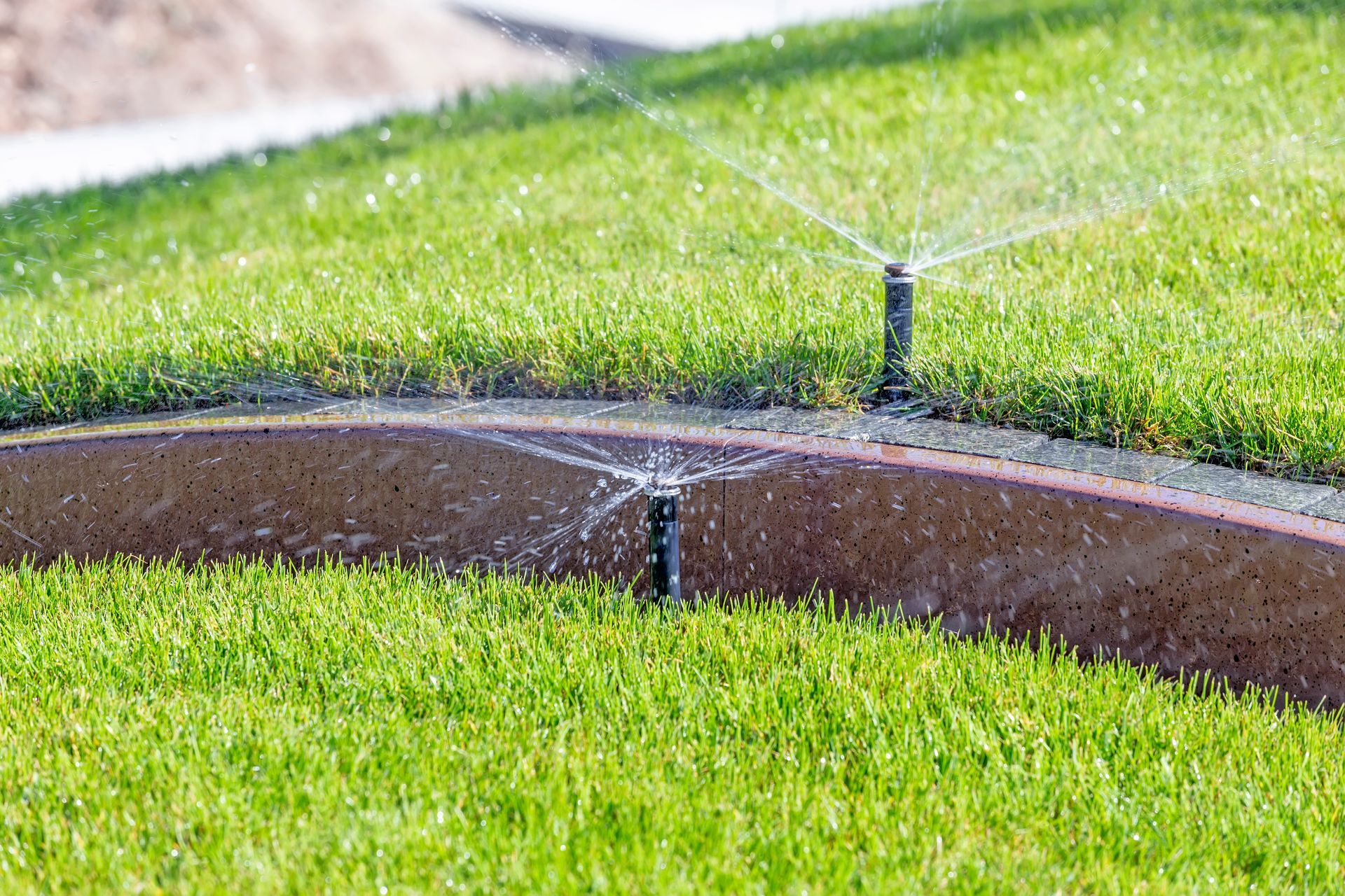 Lawn sprinkler spraying water over a green grass area beside a curved walkway