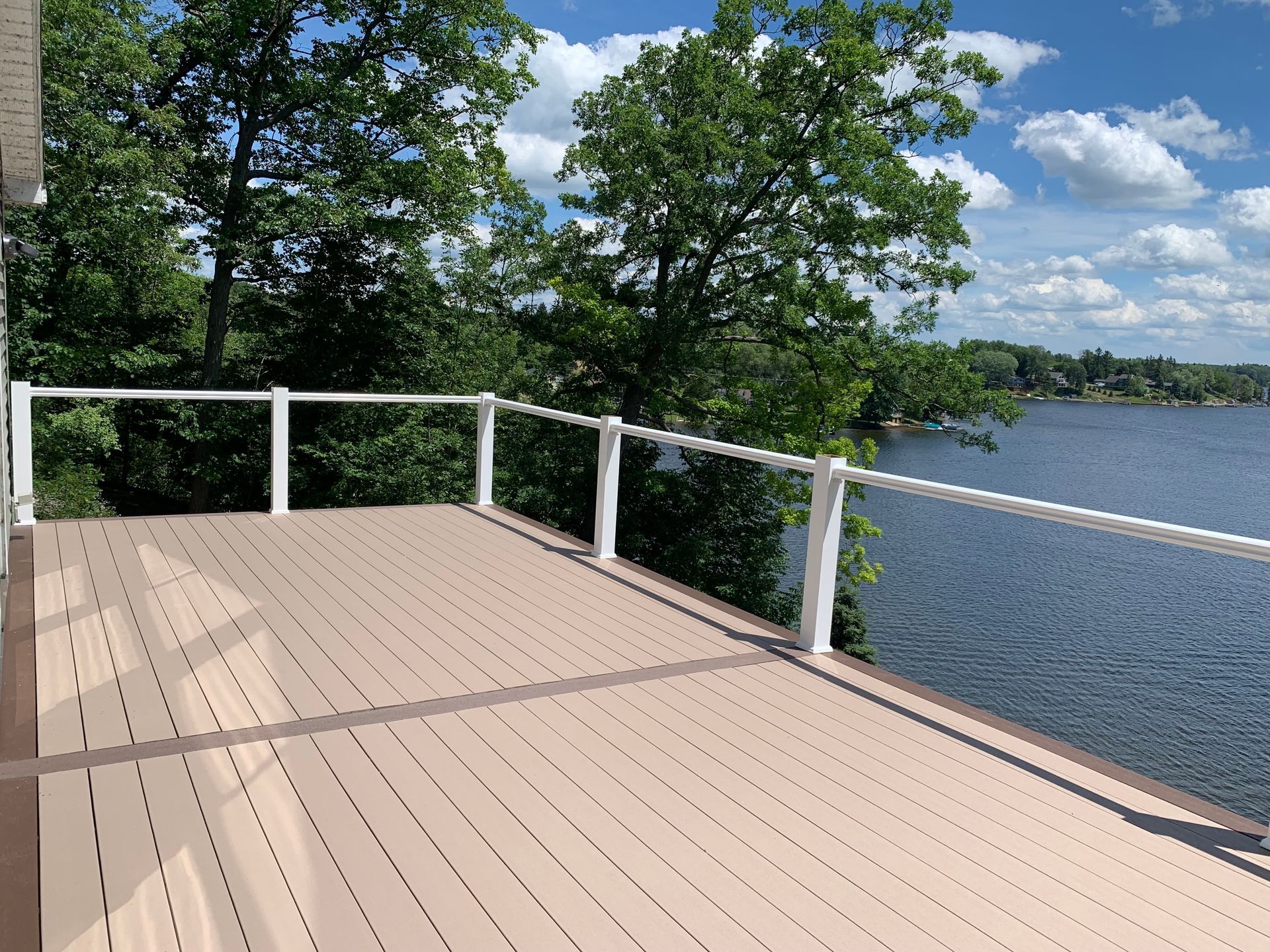 Wooden lakeside deck with white railing, overlooking calm water and trees under a partly cloudy sky