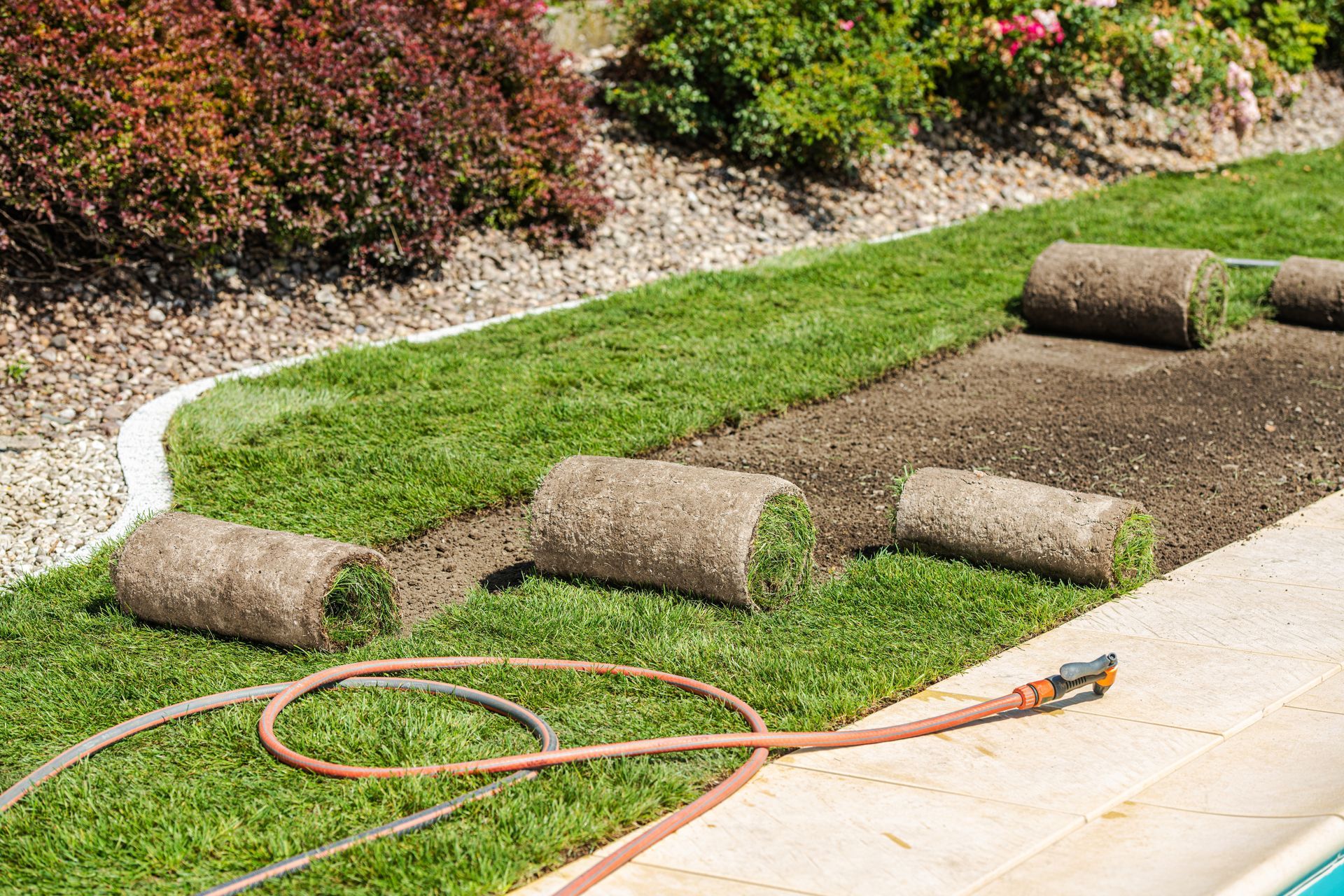 Freshly laid sod rolls on a garden bed beside a patio, with a hose and trimmed lawn nearby.
