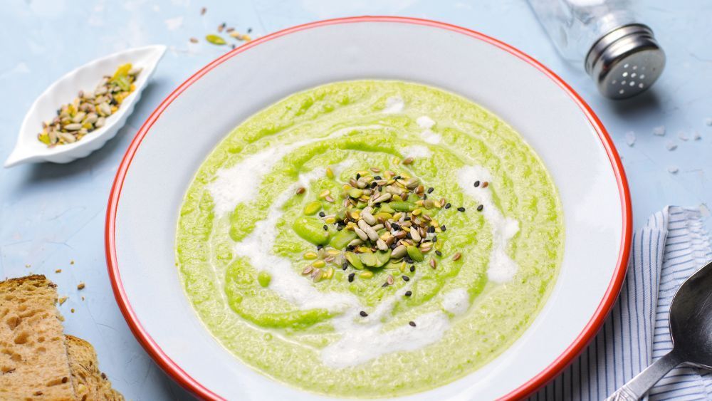 Eine Schüssel grüne Suppe mit einem Löffel und Brot auf einem Tisch.