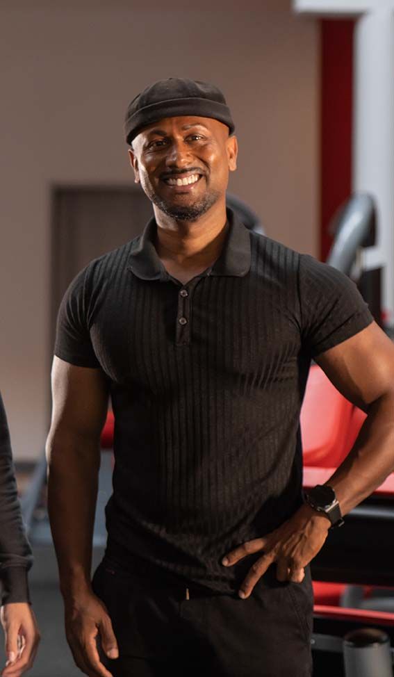 Ein Mann in einem schwarzen Hemd und Hut steht mit den Händen auf den Hüften in einem Fitnessstudio.