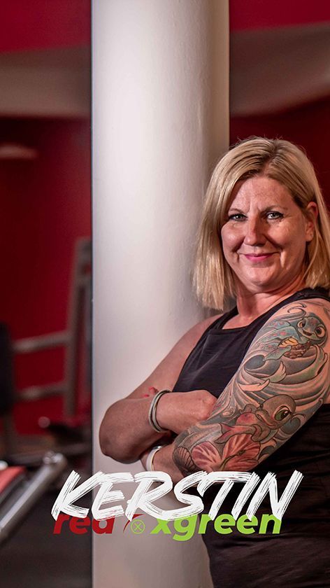 Eine Frau mit einer Tätowierung auf dem Arm steht mit verschränkten Armen in einem Fitnessstudio.