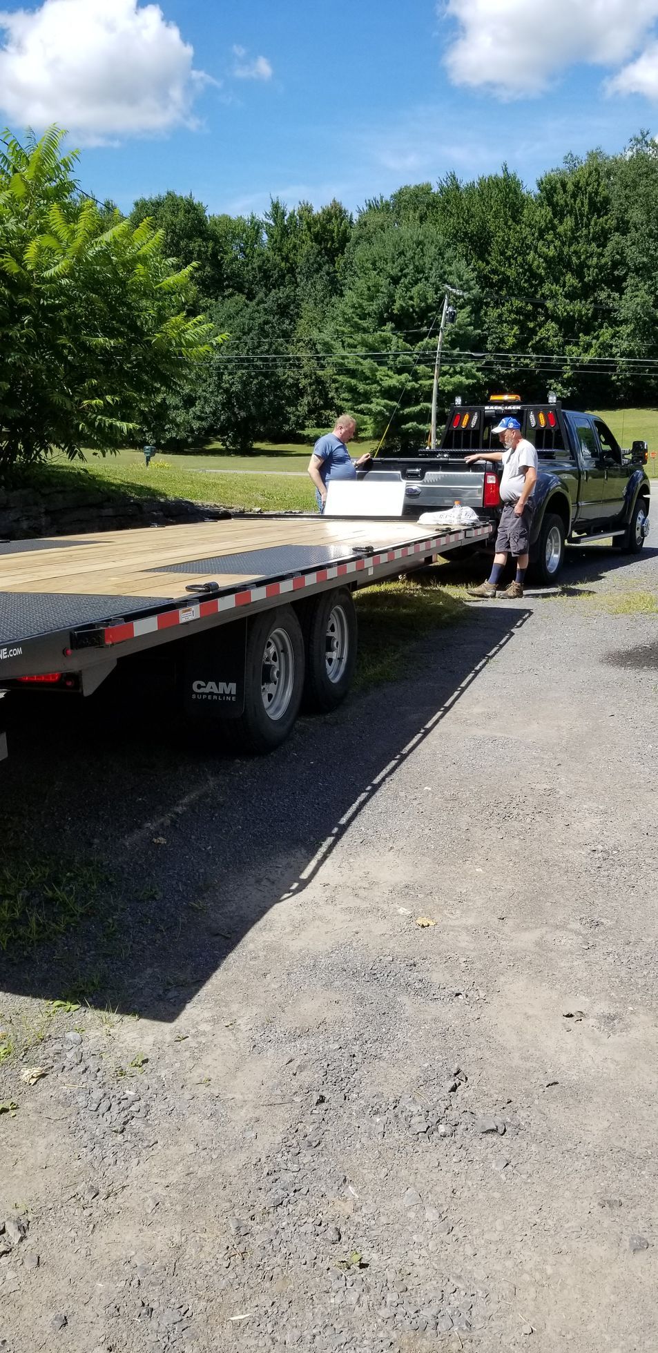 Two men loading rectangular white objects onto a flatbed trailer attached to a black truck on a gravel driveway.