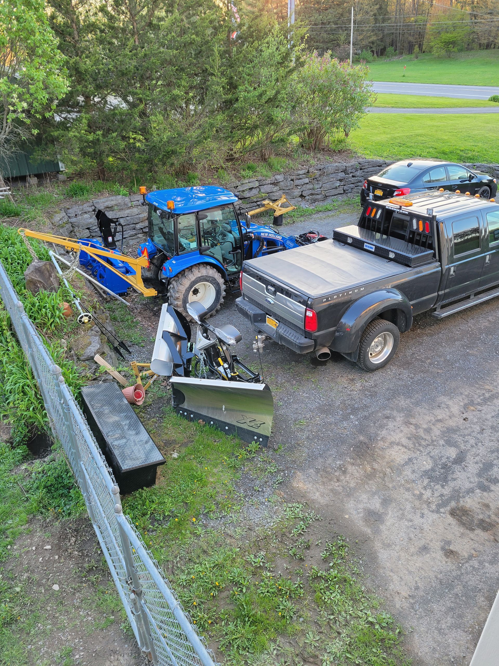 Blue tractor with snowplow next to a black truck, debris in gravel area.