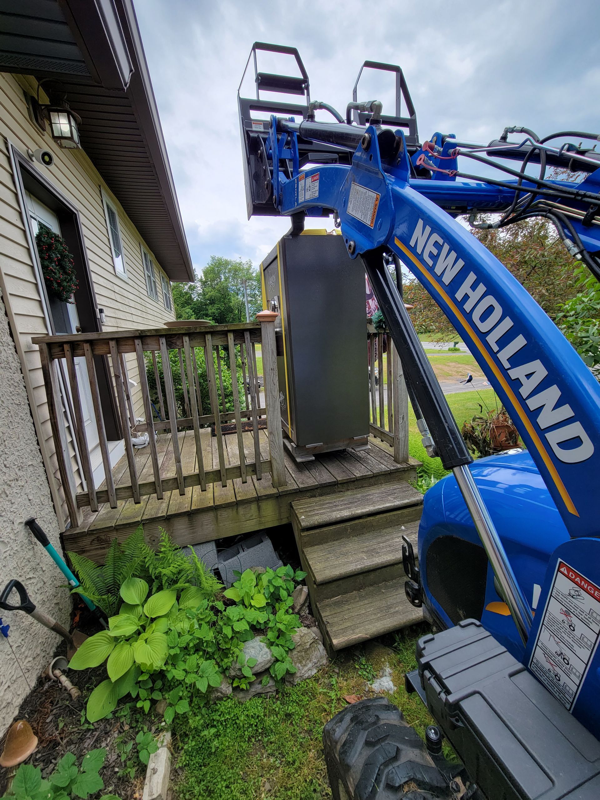 Blue New Holland telehandler lifting a dark box onto a wooden deck next to a house.
