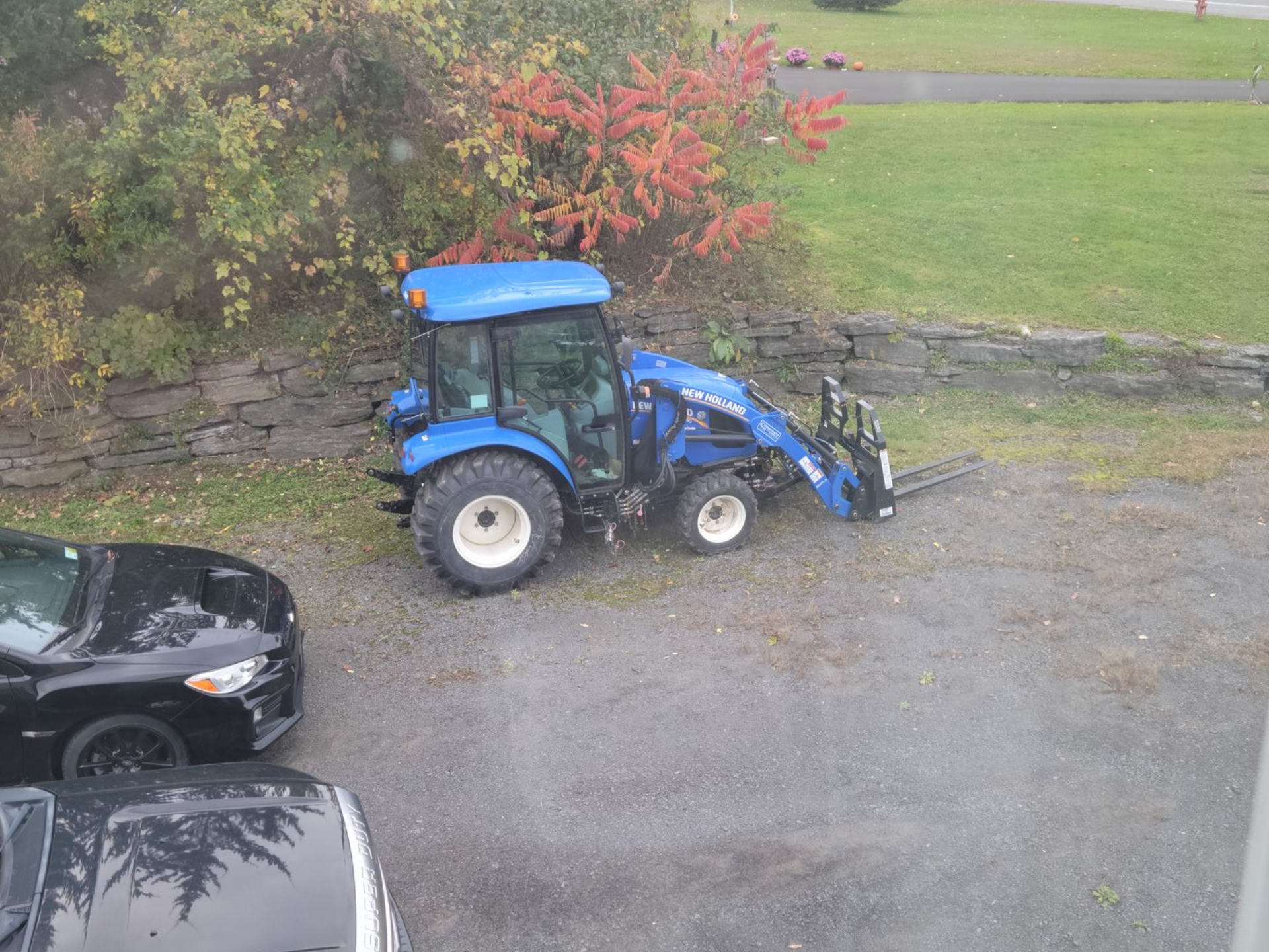 Blue tractor with a loader parked on a gravel area, next to two cars and a grassy area with fall foliage.