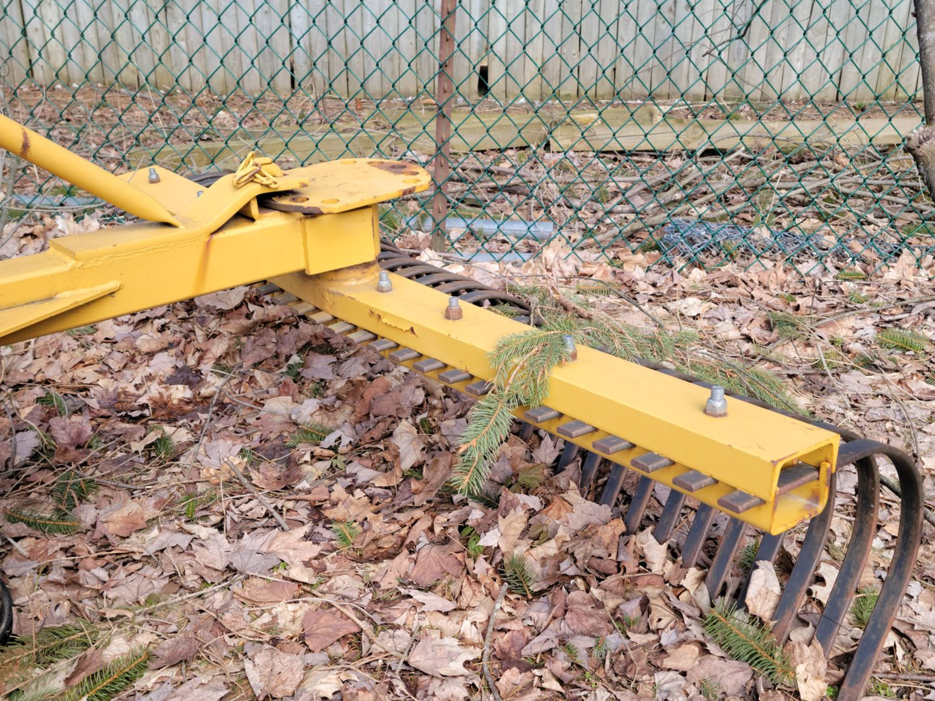 Yellow landscape rake resting in leaves; chain-link fence in background.