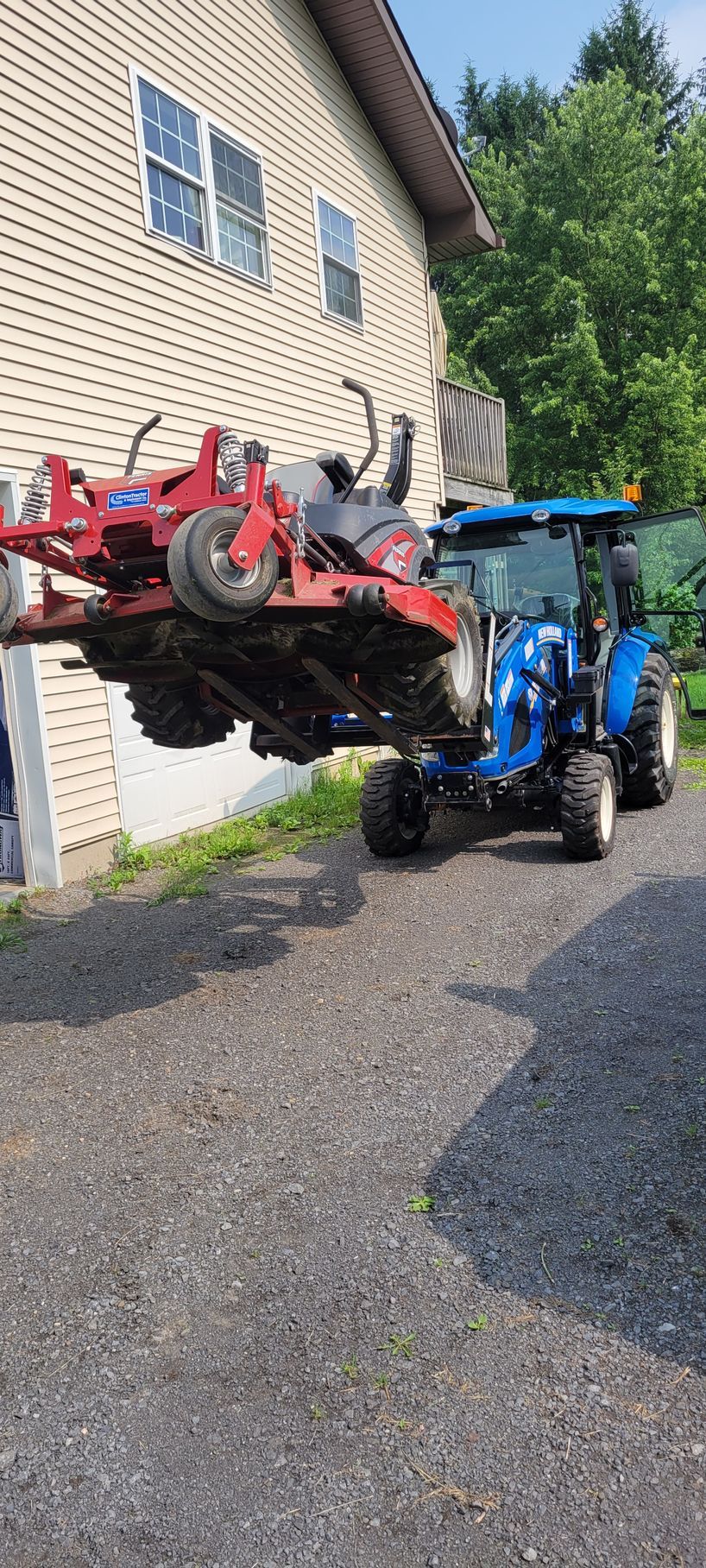 Blue tractor lifting a red zero-turn mower on a gravel driveway next to a house.