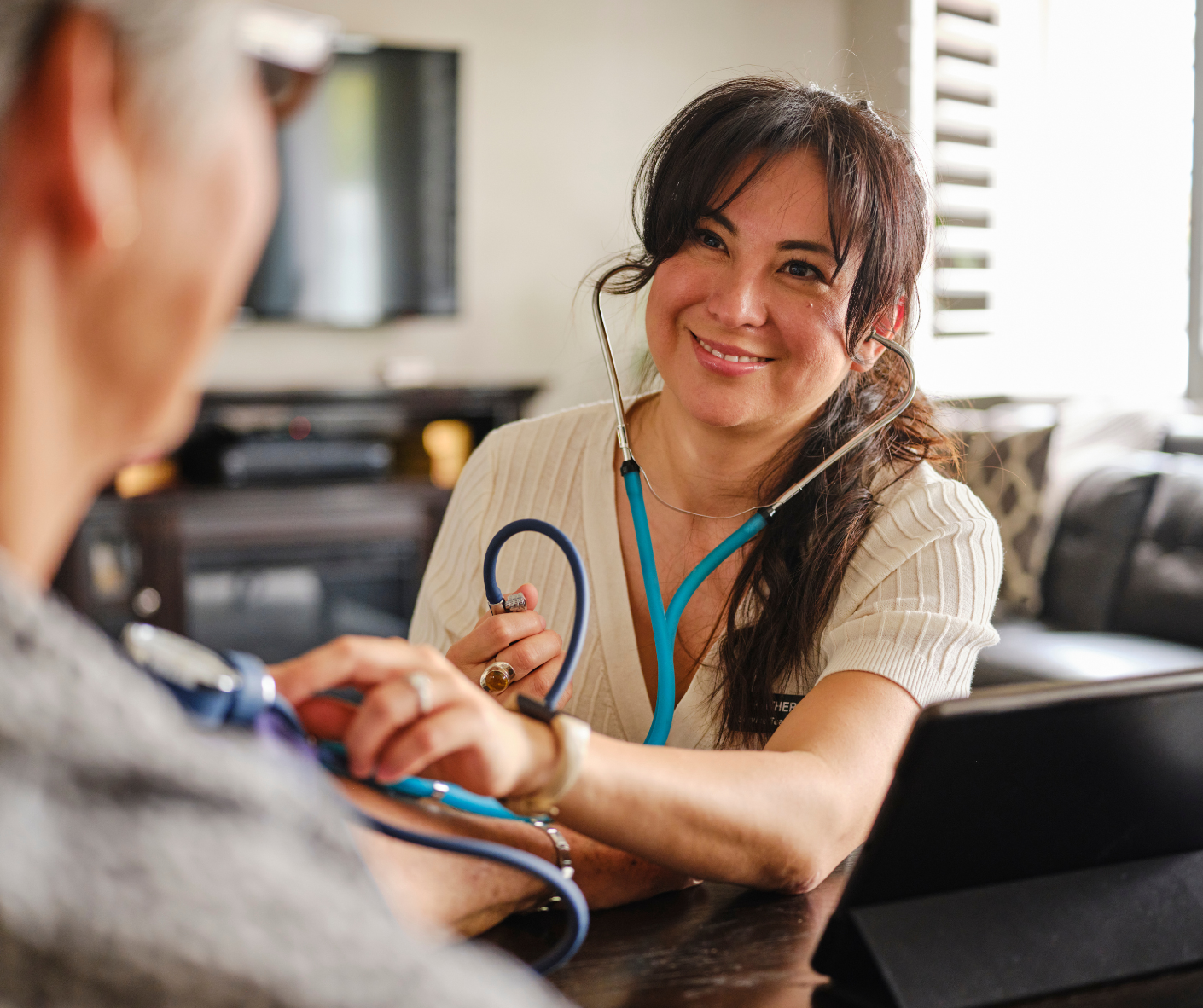 A healthcare worker smiles while checking a patient's blood pressure with a stethoscope in a home setting.