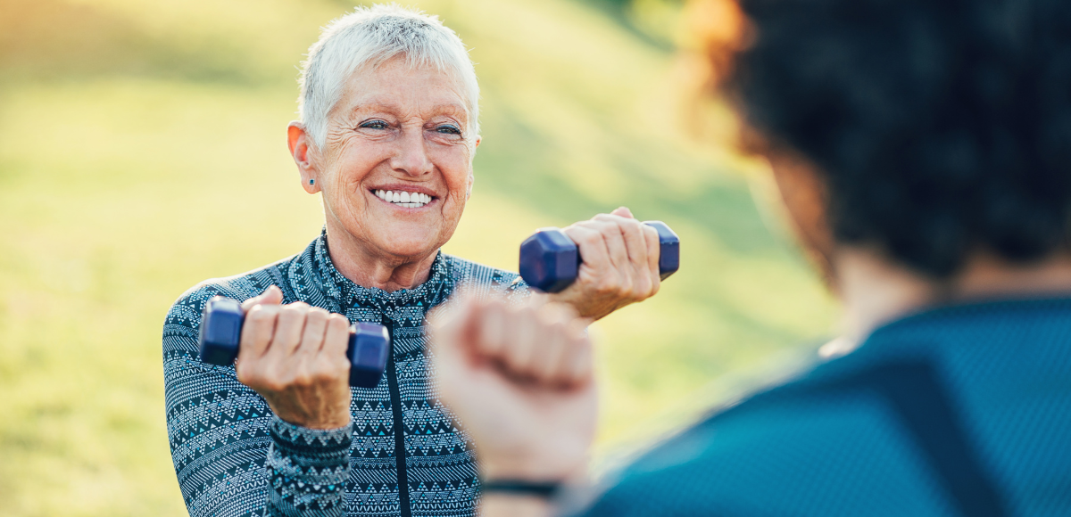 An older person exercises with dumbbells, smiling, with a person behind them. Outdoors, blurred background.