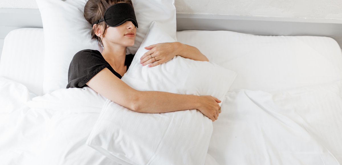 A person in a black shirt and sleep mask lies in a white bed, hugging a white pillow.