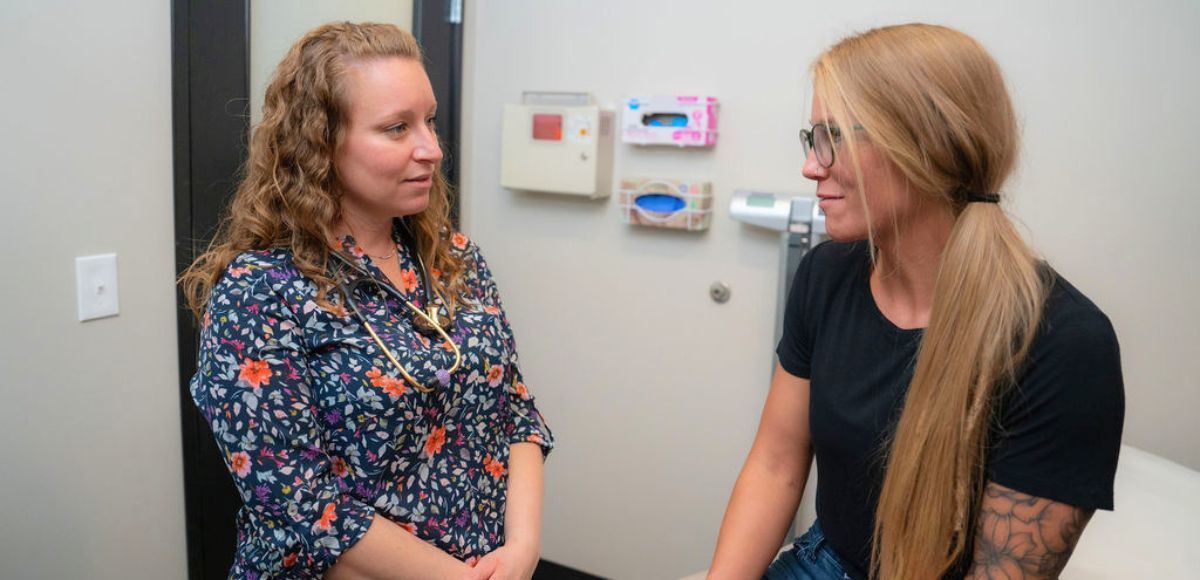 Two women talk in a medical exam room, standing near wall-mounted health monitors.
