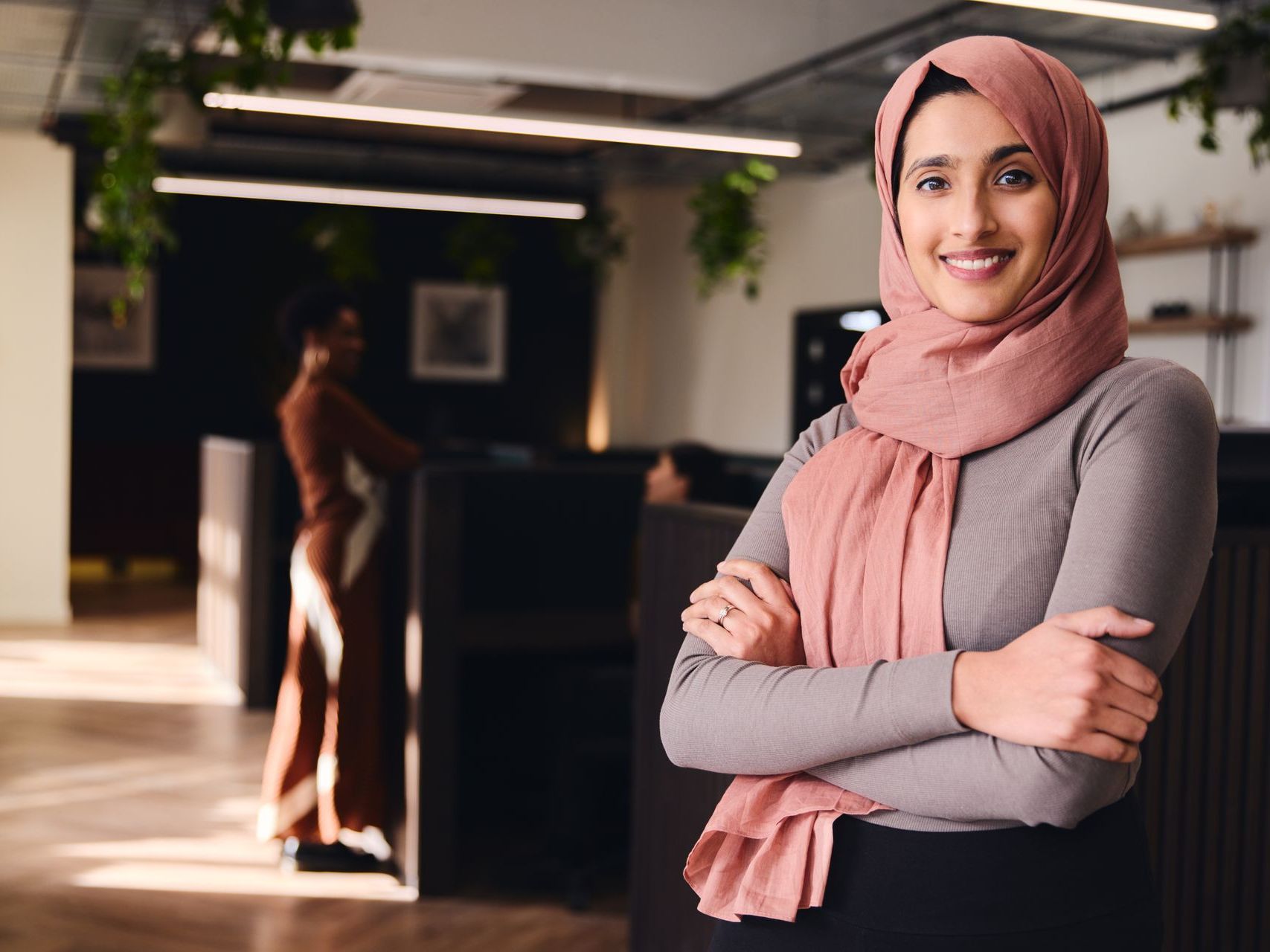A woman wearing a hijab is standing in an office with her arms crossed.