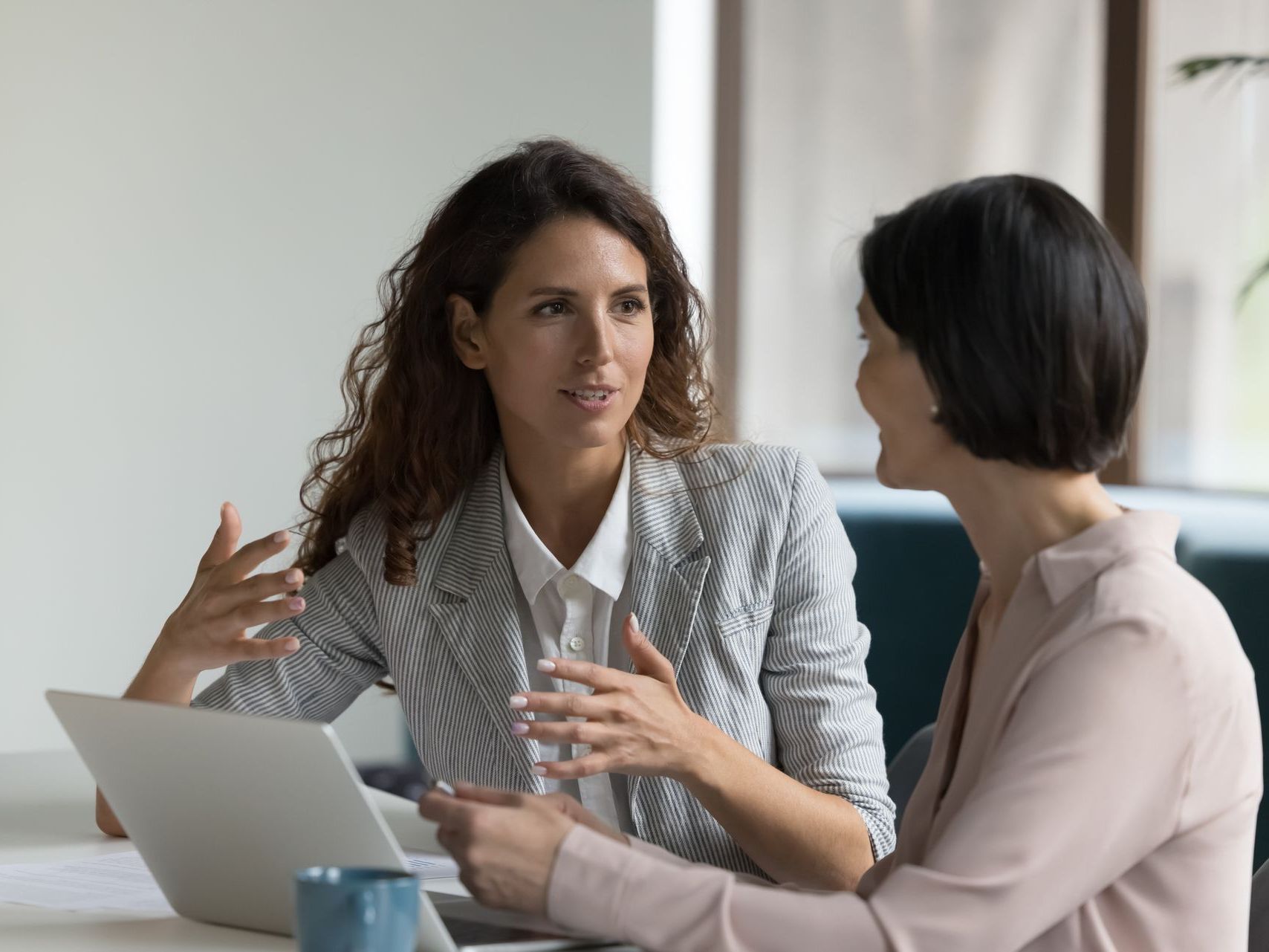 Two women are sitting at a table with a laptop and talking to each other.