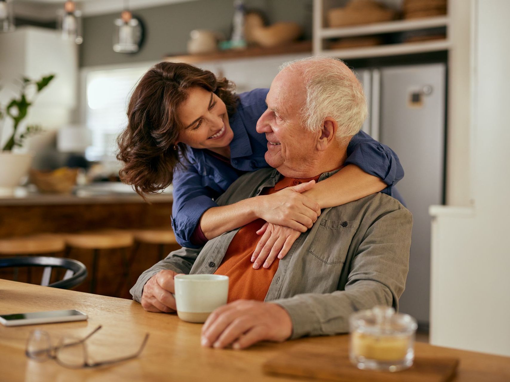 A woman is hugging an older man while sitting at a table with a cup of coffee.