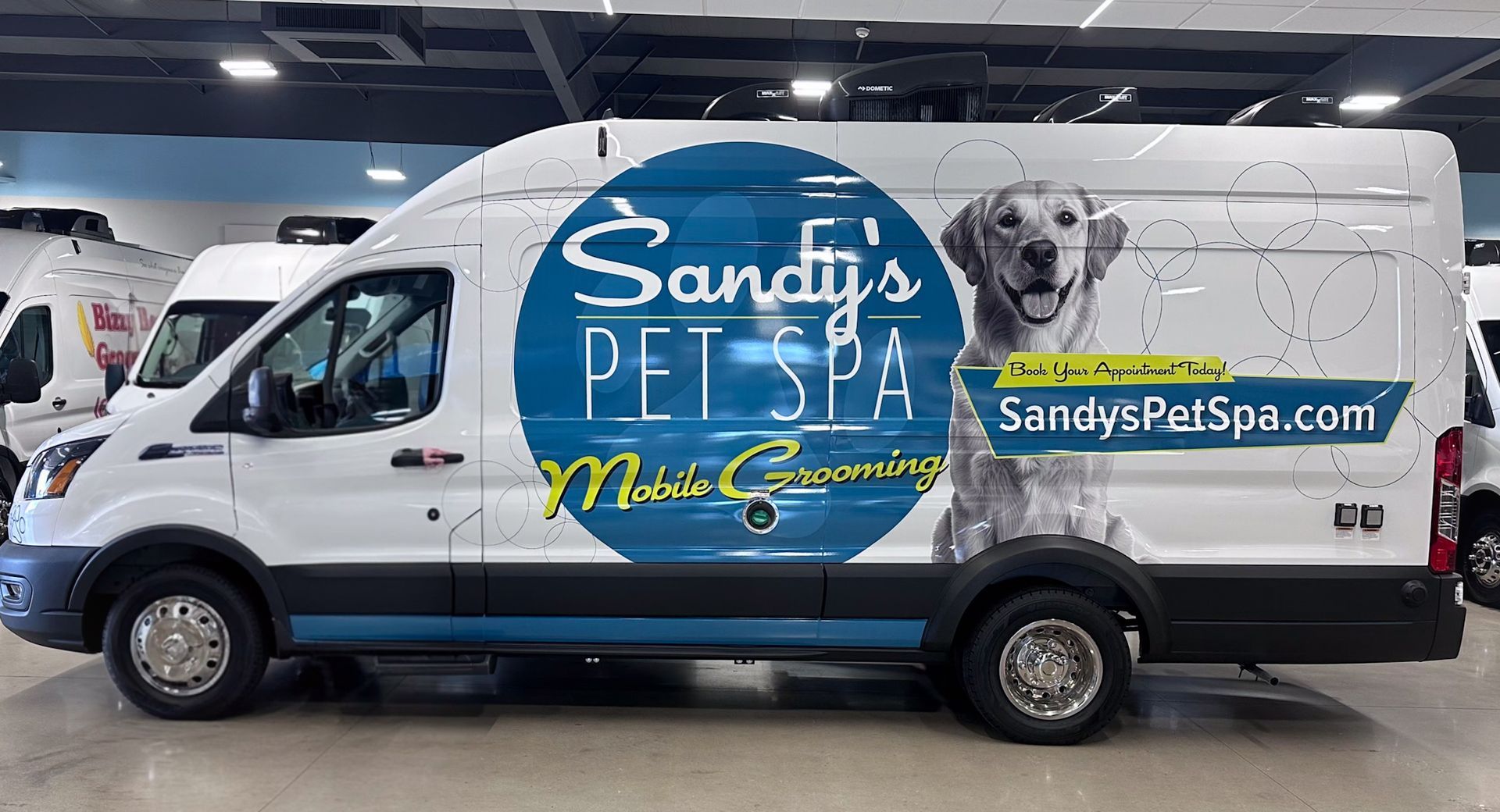White van with blue and green branding for Sandy's Pet Spa, featuring a dog illustration, parked indoors.