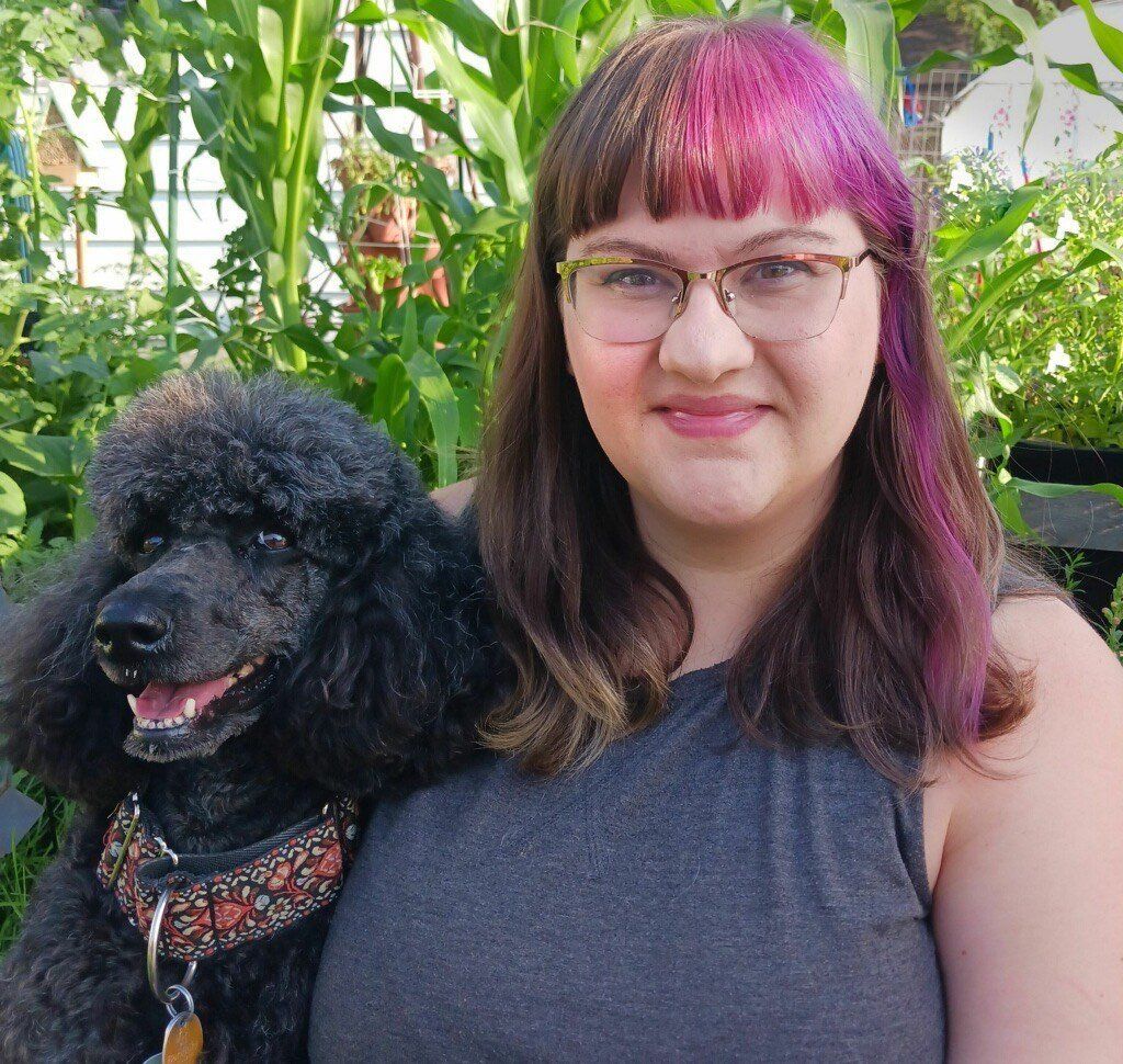 A woman with purple hair is holding a black poodle.