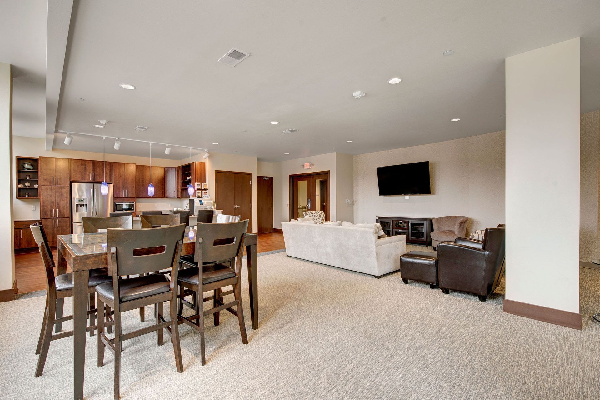 Avenir Apartments Resident Lounge. Spacious open-concept room; features a kitchen with wooden cabinets, dining table, and living area with sofas, armchairs, and a wall-mounted TV.