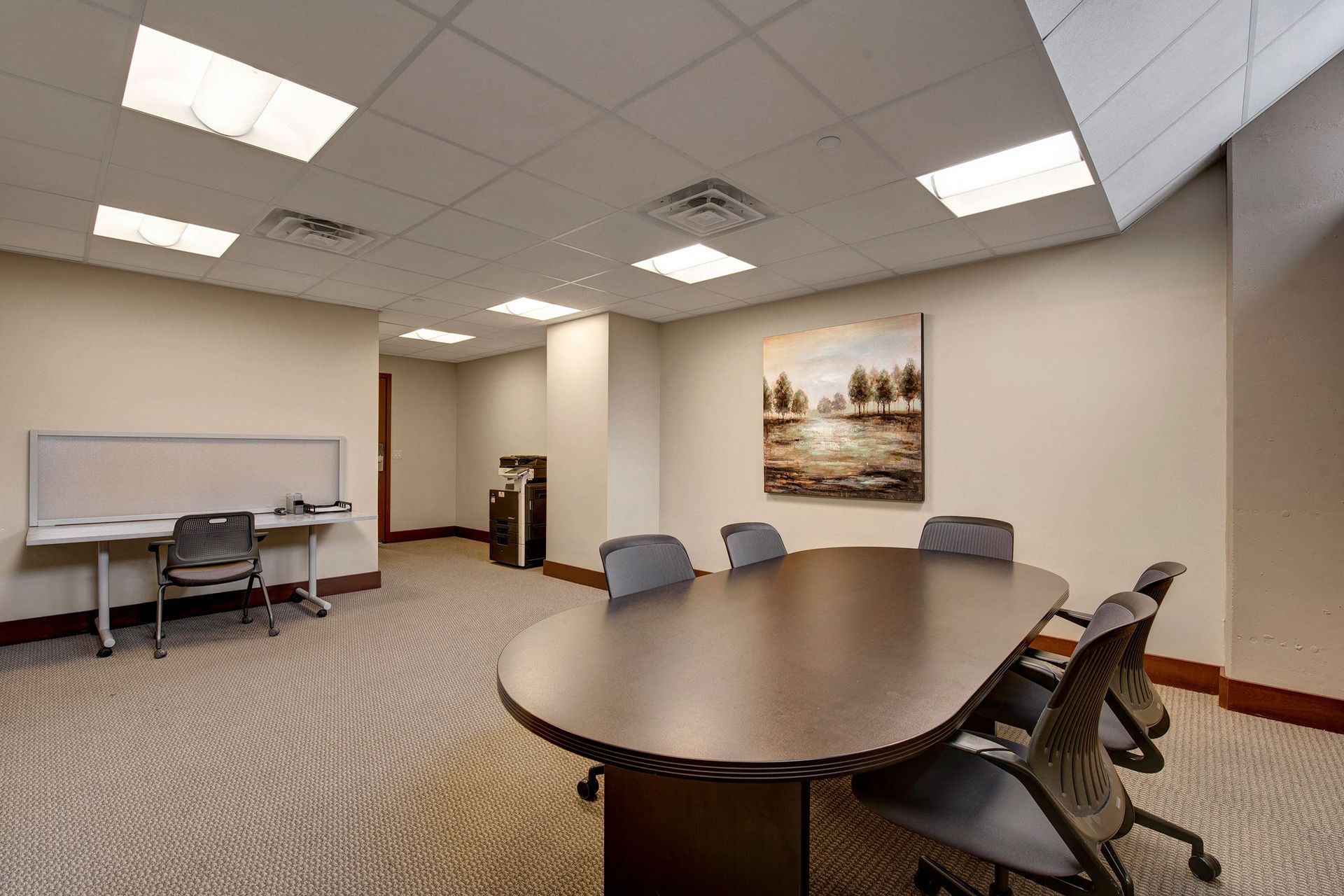 Avenir Apartments Modern conference room with oval table and six chairs on gray carpet. A landscape painting decorates the beige wall; office desk and printer in the corner.