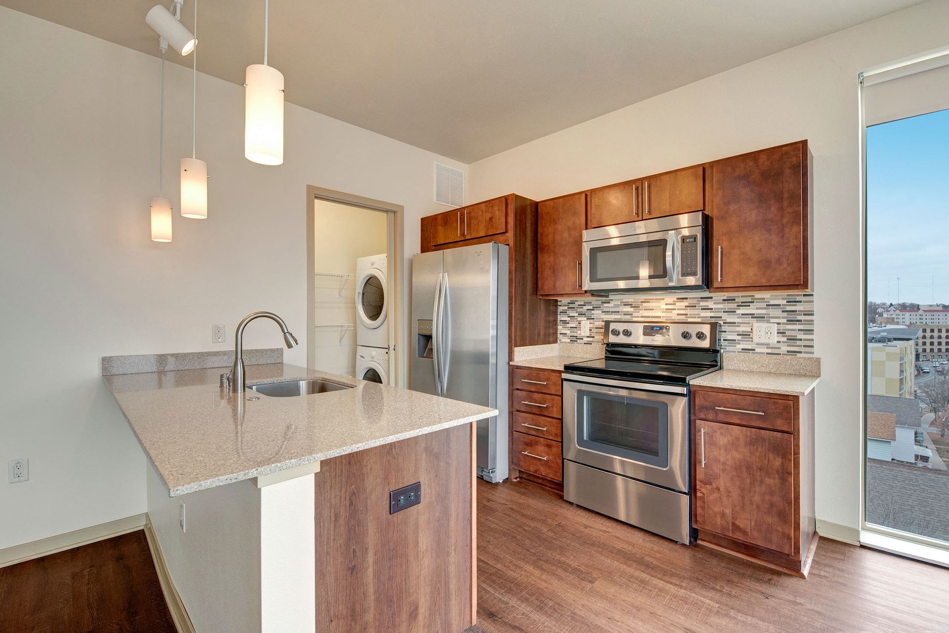 Modern kitchen at Avenie Apartments with wooden cabinets, stainless steel appliances, and a speckled countertop island. Hanging pendant lights add a warm, inviting glow.