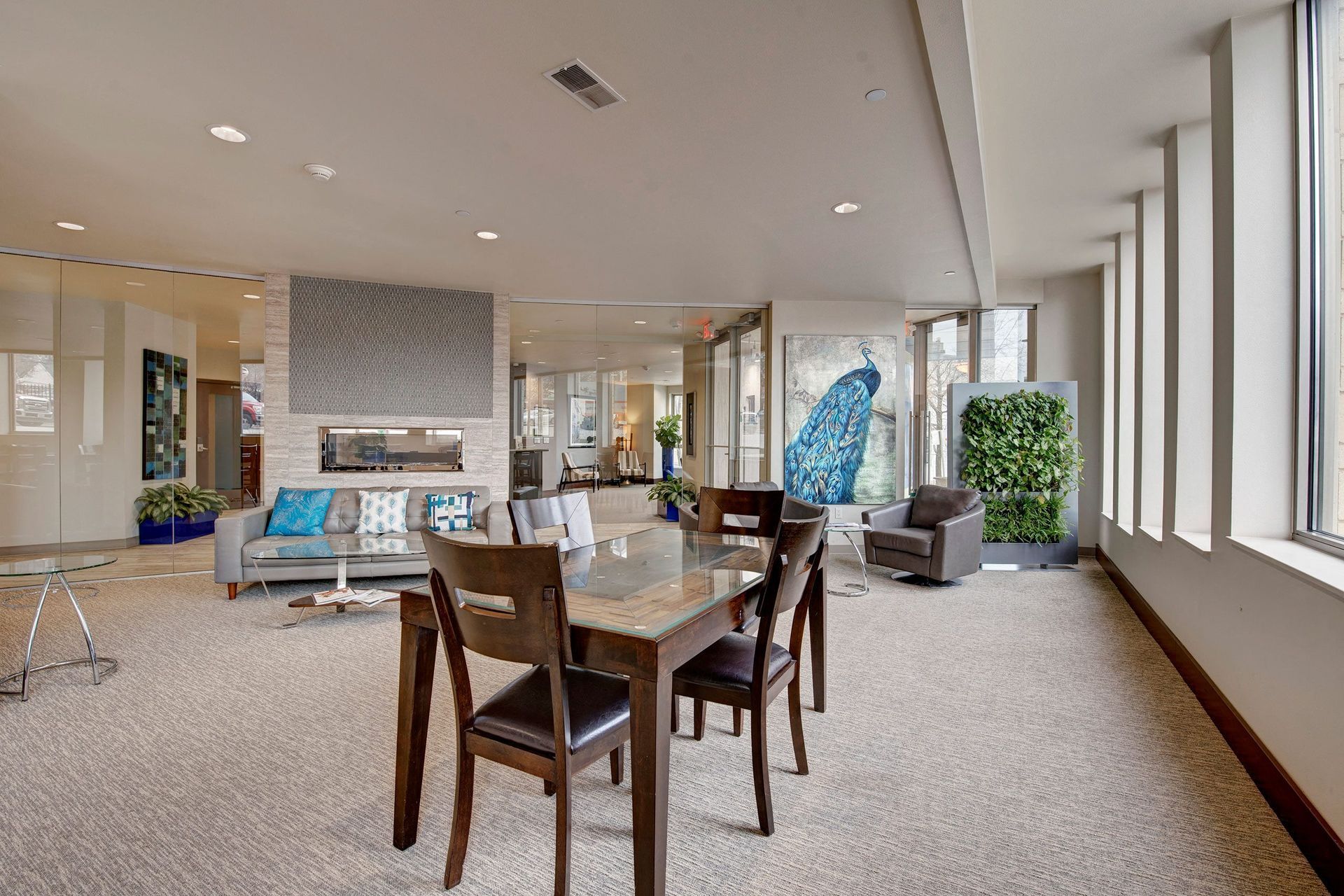 Avenir Apartments Resident lounge with gray sofas, a glass table, and dark wooden chairs. A peacock painting and vertical garden add vibrant color. Bright, airy feel.