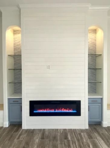 White built-in fireplace with black insert and blue flames, flanked by open shelves and gray cabinets.