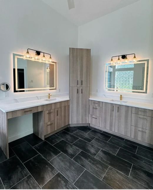 Modern bathroom with double vanities, lit mirrors, white countertops, wood cabinets, and dark tile floor.