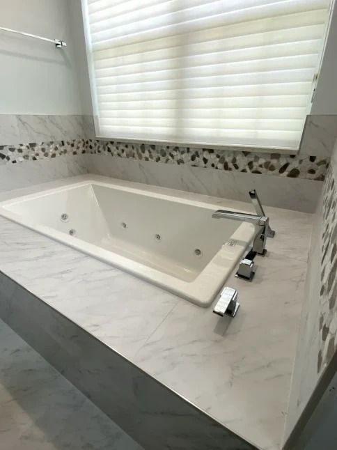 Modern bathroom with a rectangular white soaking tub set into a marble surround under a window blind.