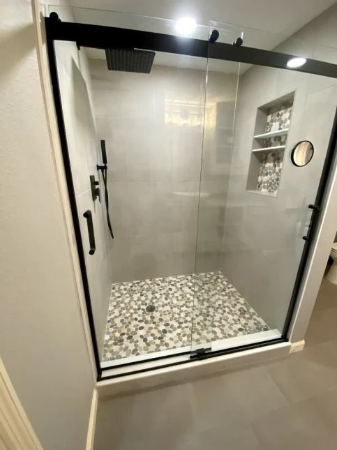 Modern glass shower stall with black frame, gray tile walls, and pebble floor in a bathroom