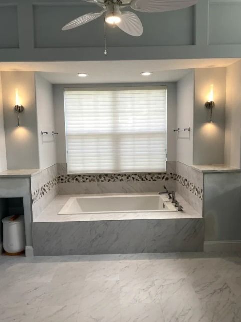 Spa-like bathroom with a marble soaking tub, gray tile walls, and a window with blinds