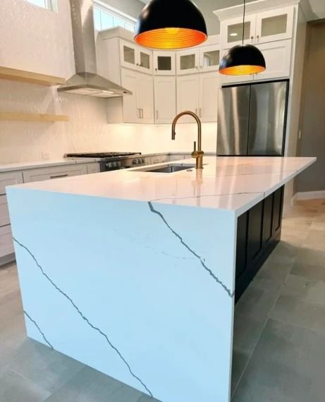 Modern white kitchen with waterfall island, brass faucet, pendant lights, and stainless steel refrigerator.