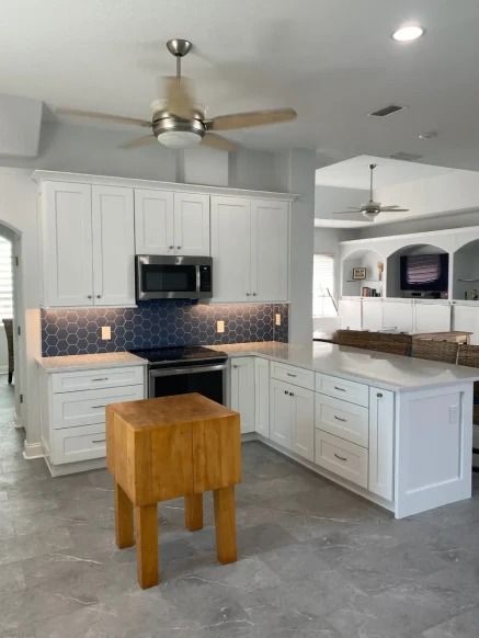 Bright white kitchen with gray tile floor, island, black appliances, and wood stool under ceiling fans