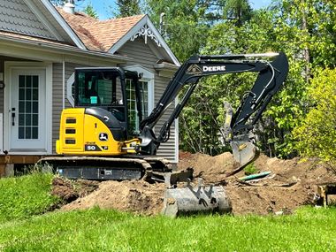 Une pelleteuse John Deere jaune et noire en train de creuser près d'une maison par une journée ensoleillée.