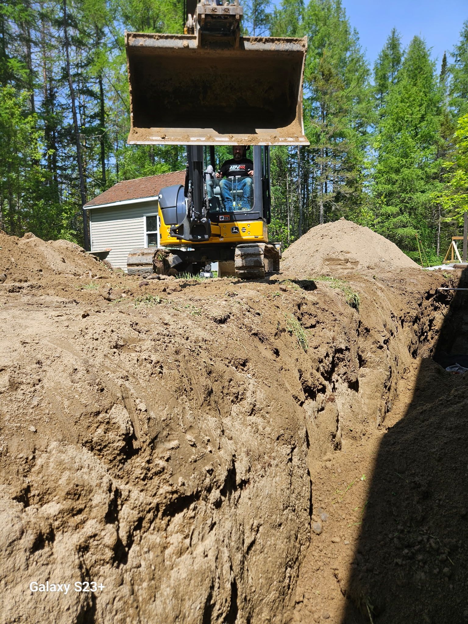 Une mini-pelle creuse une tranchée dans un jardin. Un tas de terre se dresse à l'arrière-plan, avec des arbres et une maison.