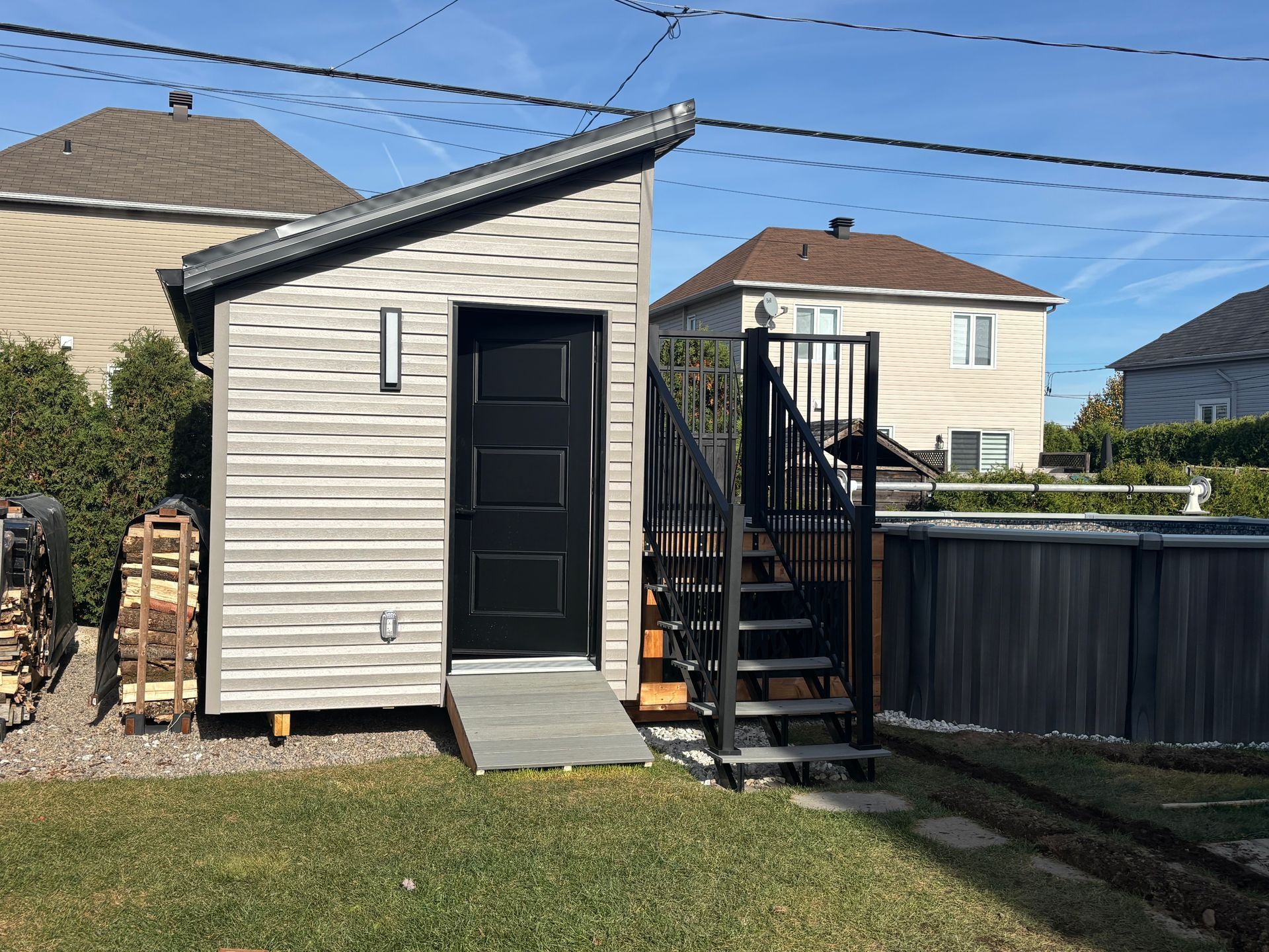 Abri de jardin avec rampe et escalier. Revêtement extérieur gris, porte noire et toit en pente.