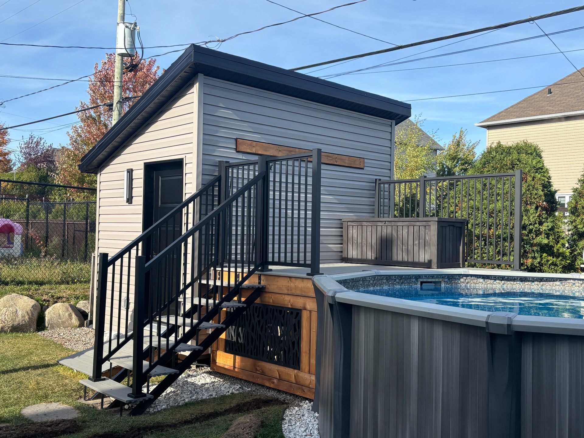 Petit cabanon avec rambarde noire, escalier et terrasse donnant sur une piscine.